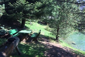 A picnic area with benches and a table, surrounded by trees and a body of water. - Glacier Chalet by the pond (Flattach)