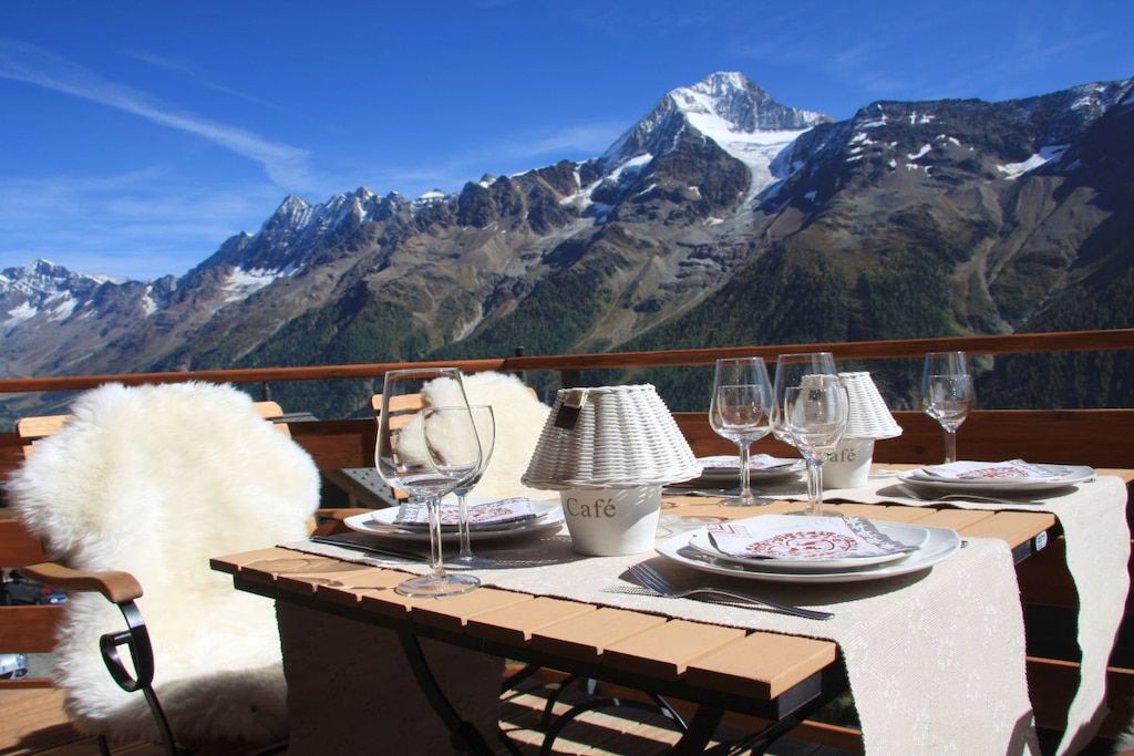 Ein Esstisch für vier Personen, gedeckt mit Tischdecke, mit Blick auf die Berge und einem weißen, flauschigen Stuhl.