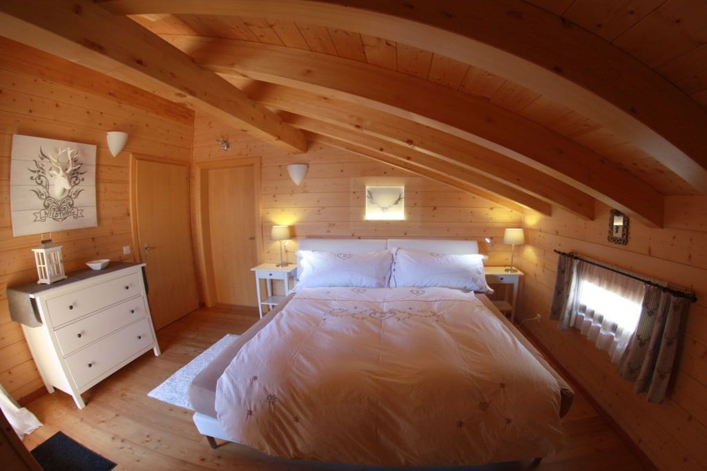 Ein Schlafzimmer mit Holzbalkendecke, einem Bett, einem Schminktisch, Nachttischen und einem Fenster mit Vorhängen.