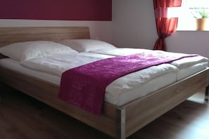 Een eenpersoonsbed met houten frame en witte beddengoed, een paars plaid en een raam met rode gordijnen.