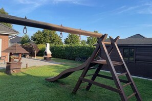 A wooden swing set in a backyard with a brick house and a barbecue grill in the background. - Holiday home Stein in the Spreewald Lübbenau (Lübbenau)