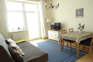 Ein Zimmer mit einem Bett, einem Fernseher, einem Esstisch mit Stühlen, einem Fenster mit Vorhängen und einem Bild an der Wand.