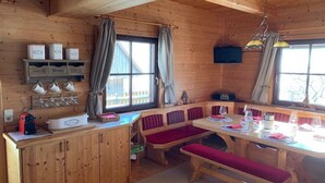 A wooden cabin interior with a dining table set for two, a bench, a shelf with mugs, and a television mounted on the wall. - Kogljahrerhütte || Prebl (Prebl)