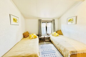 Ein kleines, gut beleuchtetes Zimmer mit zwei Einzelbetten, einem kleinen Tisch und einem Fenster mit Vorhängen.