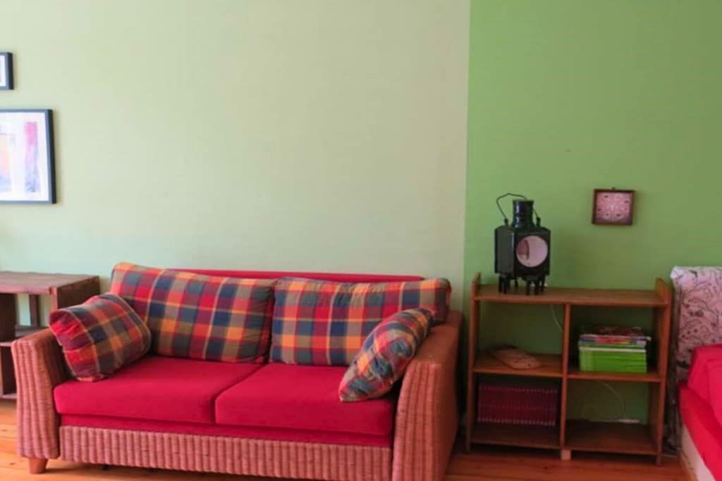 Ein rotes Sofa mit karierten Kissen, ein Holztisch, eine grüne Wand und ein Regal mit Büchern und einem Radio.