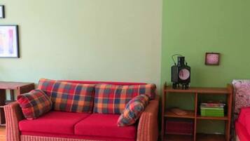 Ein rotes Sofa mit karierten Kissen, ein Holztisch, eine grüne Wand und ein Regal mit Büchern und einem Radio.