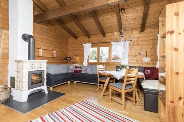 A cozy cabin interior with a wood-burning stove, a sofa, a dining table, and a window with curtains.