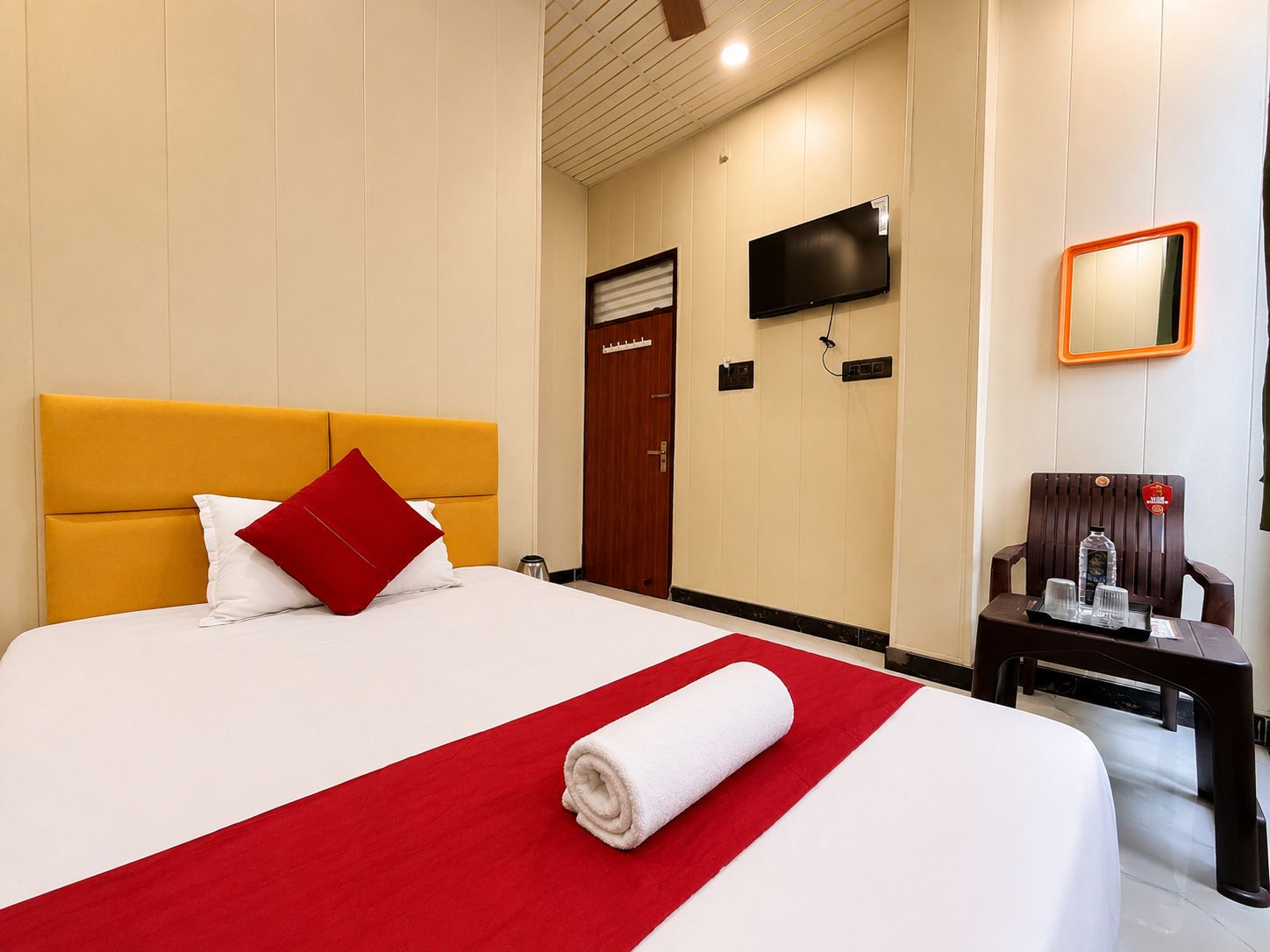 Ein Hotelzimmer mit einem Bett, einer roten und weißen Tagesdecke, einem weißen Handtuch, einem hölzernen Nachttisch, einem Spiegel und einem Fernseher an der Wand.