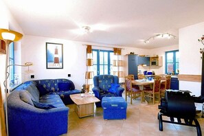 A living room with a sofa, armchair, ottoman, and a dining area with chairs and a table. - Holiday home in Glowe with swimming pool (Glowe)