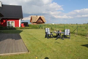 A wooden deck with a table and chairs, a red building, and a large house with a sloped roof in the background. - Holiday home in Lancken with swimming pool (Lancken)