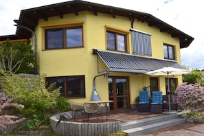 Una casa de dos pisos con fachada amarilla, ventanas grandes y un porche cubierto con mobiliario de exterior.