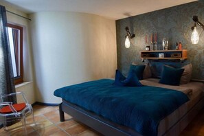 Un dormitorio con cama, una silla, un estante con objetos y una pared con estampado.
