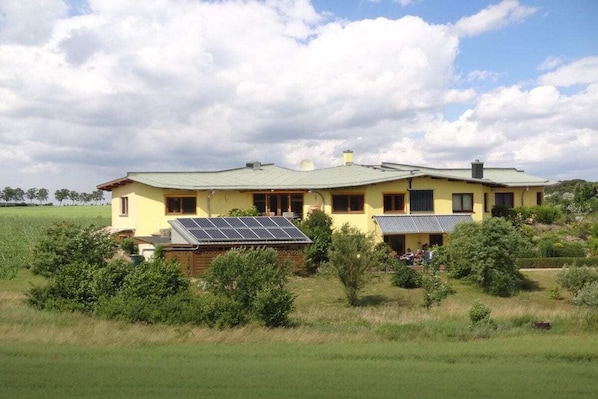 Una casa de dos pisos con techo verde, rodeada de follaje y un campo.