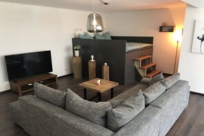Una sala moderna con un sofá modular, un televisor de pantalla plana, una mesa de centro de madera, una mesita auxiliar pequeña y una cama litera con escalera.