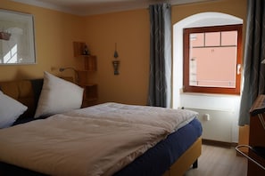 Un dormitorio con cama, ventana con cortinas, radiador y estante de pared con un objeto pequeño.