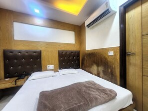 A hotel room with a bed, a wooden headboard, a bedside table with a telephone, and an air conditioning unit. - Oly Hotel Cottage lord Krishna  (New Delhi)