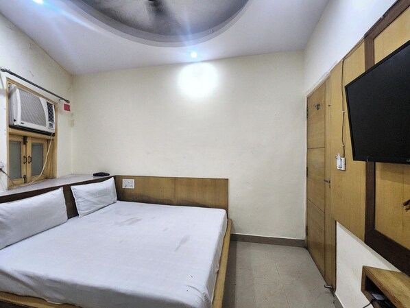 Una habitación pequeña con cama individual, cabecera de madera, televisor montado en la pared y ventana con aire acondicionado.