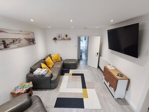 Un salon moderne avec un canapé gris, des coussins jaunes, un pouf noir et un grand téléviseur à écran plat.