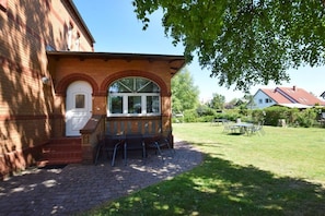 Ein Backsteinhaus mit weißer Tür und Fenstern, ein gepflasterter Bereich mit Bank und Stühlen sowie eine Rasenfläche mit Tisch und Stühlen.