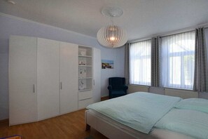 Ein Schlafzimmer mit Bett, Schrank, Regalen, einem Sessel und einem Fenster mit Vorhängen.