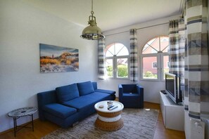 Sala de estar com um sofá azul, uma mesa redonda pequena, uma televisão e um quadro na parede.