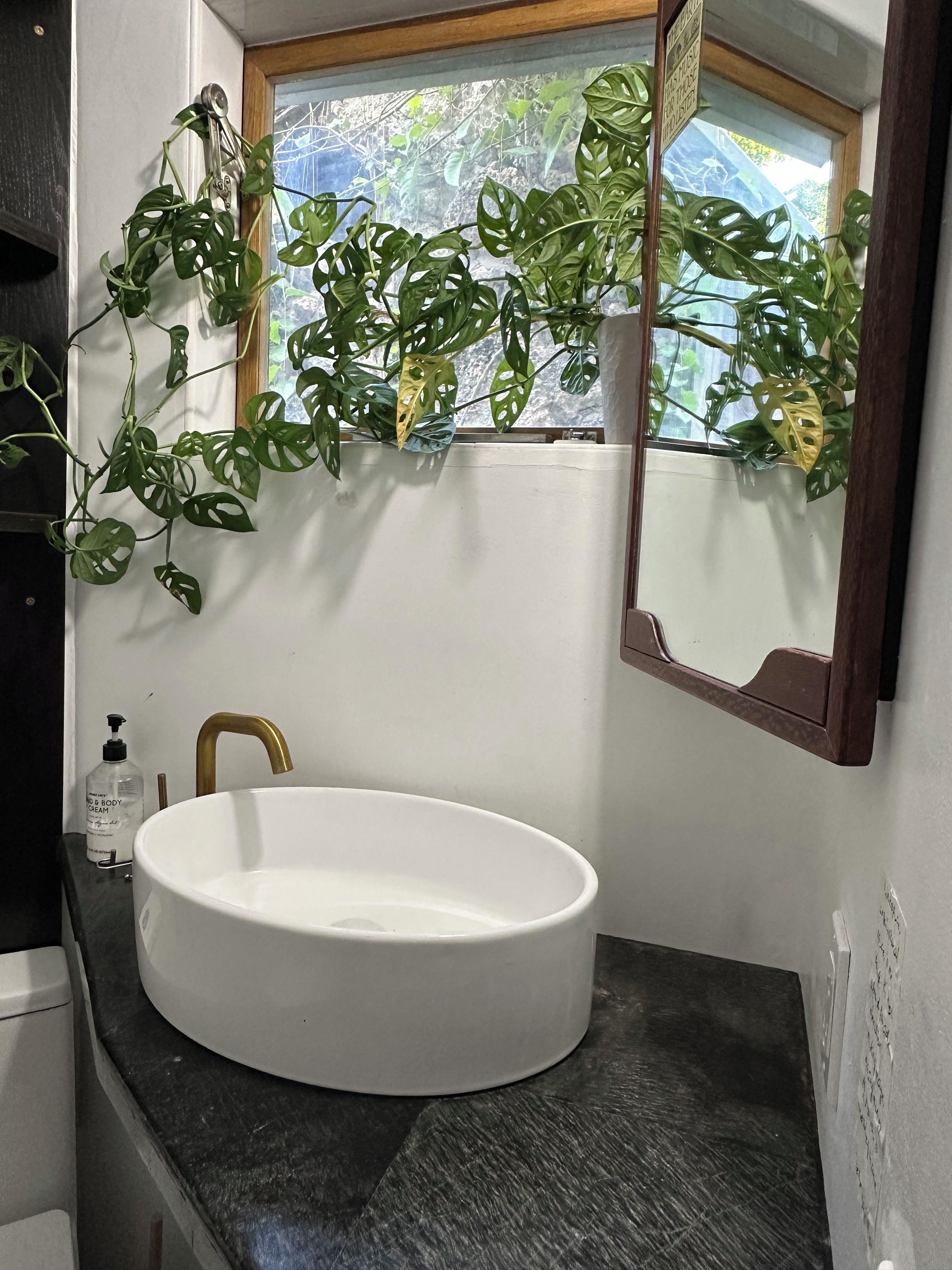 Un bagno con lavandino bianco, rubinetto dorato, specchio e finestra vista verde.
