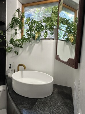 Un bagno con lavandino bianco, rubinetto dorato, specchio e finestra vista verde.