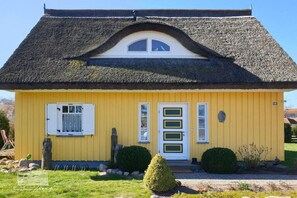 Ein gelbes Gebäude mit einem Reetdach, einer weißen Tür mit grünen Akzenten und einem kleinen Fenster.
