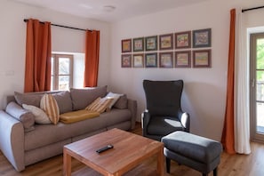 Una sala de estar con sofá, sillón, reposapiés y una mesa de centro. Hay cortinas, un ventanal y cuadros en la pared.
