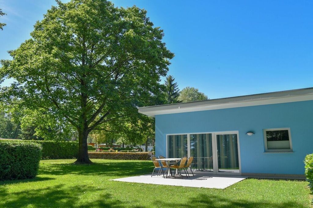 Un edificio moderno con fachada azul, un patio con mesa y sillas, y un árbol grande en primer plano.