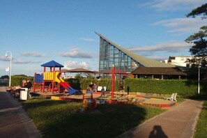 Ein Spielplatz mit Rutschen und Schaukeln, ein Gebäude mit einem markanten Dach und ein klarer Himmel.