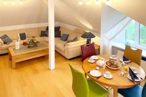 A cozy living area with a sofa and a coffee table, a dining table set for breakfast, and a window with curtains. - Captain's Garden W13 "Roth" (Prerow)