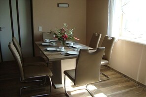 A dining area with a table set for four, surrounded by four chairs, a vase with flowers, and a window with curtains. - Holiday home with 2 bedrooms (Rosenthal-Bielatal OT Bielatal)