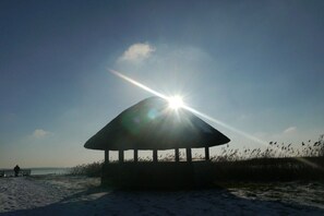A beach with a gazebo structure, a person sitting on a bench, and the sun shining through the sky. - House "Am Brunnen" Where 7 (Ostseebad Prerow)
