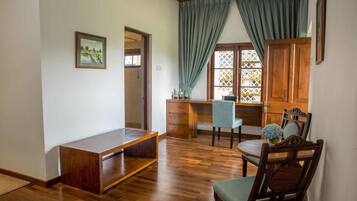Una habitación con un banco de madera, un escritorio y una silla. Se ve una ventana con cortinas y vistas al verdor exterior.