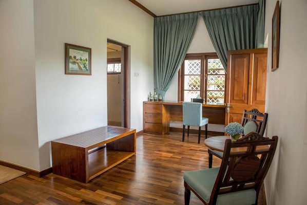 A room with a wooden bench, a desk, and a chair. There is a window with curtains and a view of greenery outside.