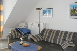 Een woonkamer met een bank, een bijzettafeltje met een fles en schaal, een vloerlamp en twee ingelijste foto's aan de muur.