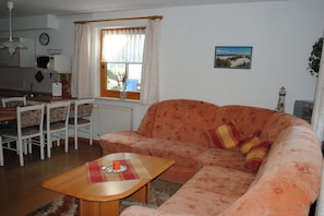A living room with a sofa, a coffee table, a dining table with chairs, a window with curtains, and a kitchen area in the background. - Terraced house with 2 bedrooms (Ostseebad Prerow)