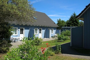 A blue building with a sloped roof, a paved patio area, and a garden with red flowers. - Terraced house with 2 bedrooms (Ostseebad Prerow)