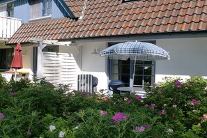 Ein Haus mit Ziegeldach, einer Terrasse mit Tisch und Stühlen sowie ein Garten mit Blumen.