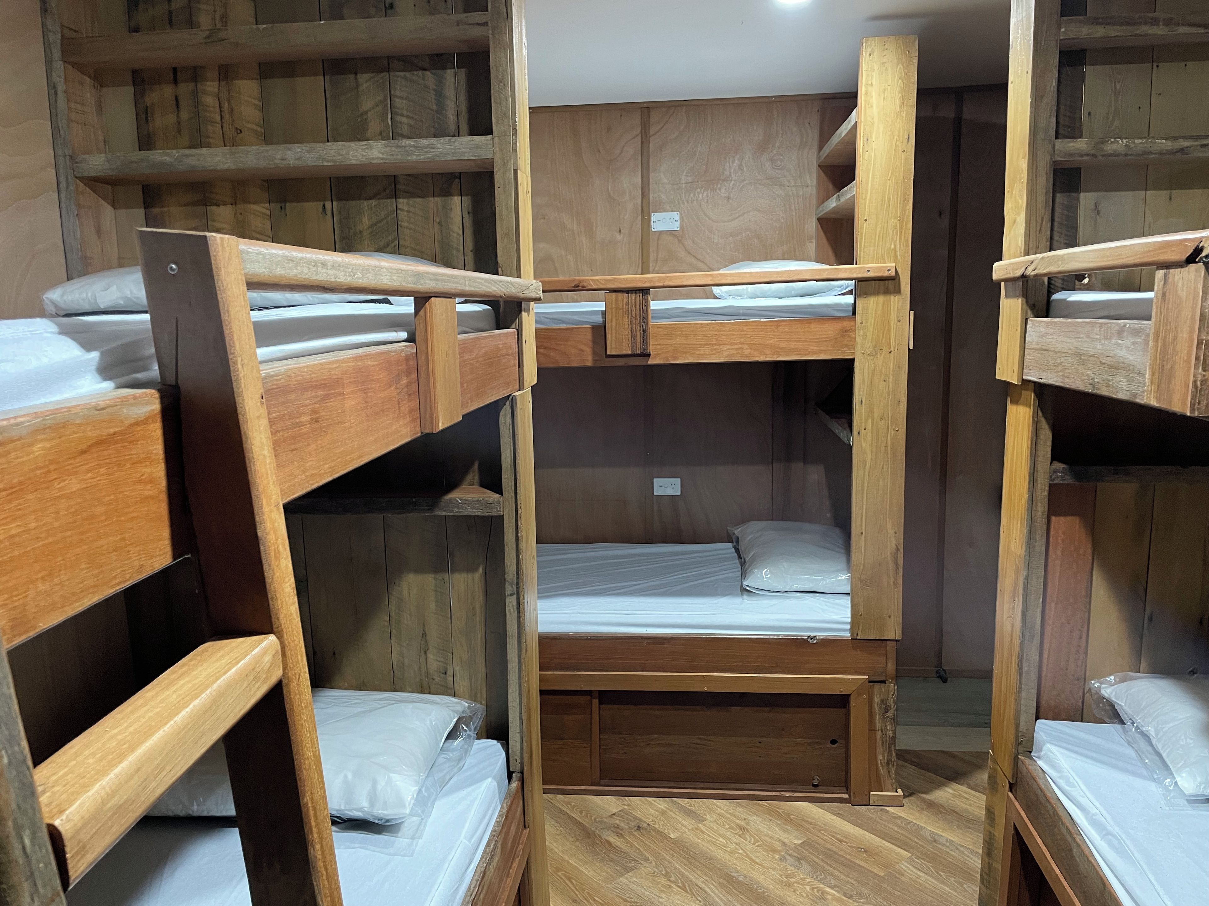 Ein Schlafraum mit Etagenbetten, Holzwänden und Holzdielen, einem Einzelbett in der Mitte und einer kleinen Bank.