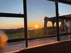 En vy genom ett fönster med en träveranda, en solnedgång och ett avlägset landskap.