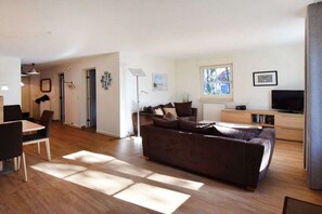 A modern living room with a brown sofa, a dining table with chairs, a flat-screen TV, and a window providing natural light. - Holiday home with 3 bedrooms (Ostseebad Prerow)