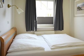 Enkeltseng med hvide sengetøj i et rum med et vindue, en lampe på væggen og en radiator.