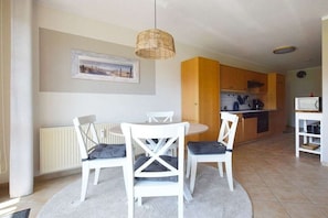 A dining area with a round table and four chairs, a kitchen in the background, and a single hanging light fixture. - Buchengrund W 1 || Ostseebad Prerow (Ostseebad Prerow)