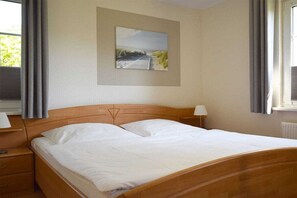 A single bed with a wooden headboard, a bedside table, a lamp, and a painting of a beach scene on the wall. - Buchengrund W 1 || Ostseebad Prerow (Ostseebad Prerow)
