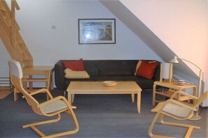 Ein Zimmer mit einem Holztisch, zwei Stühlen, einer Couch mit Kissen, einer Lampe und einem gerahmten Bild an der Wand.