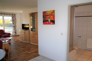 A living room with a television, a dining area with a table and chairs, a kitchen area with cabinets, and a bathroom with a shower. - House "Am Brunnen" W.2 (Ostseebad Prerow)