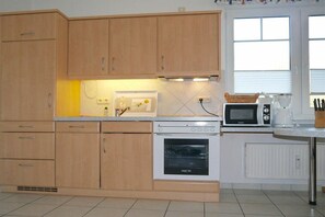 A kitchen with wooden cabinets, a built-in oven, a microwave, and a window with blinds. - House "Am Brunnen" W.2 (Ostseebad Prerow)