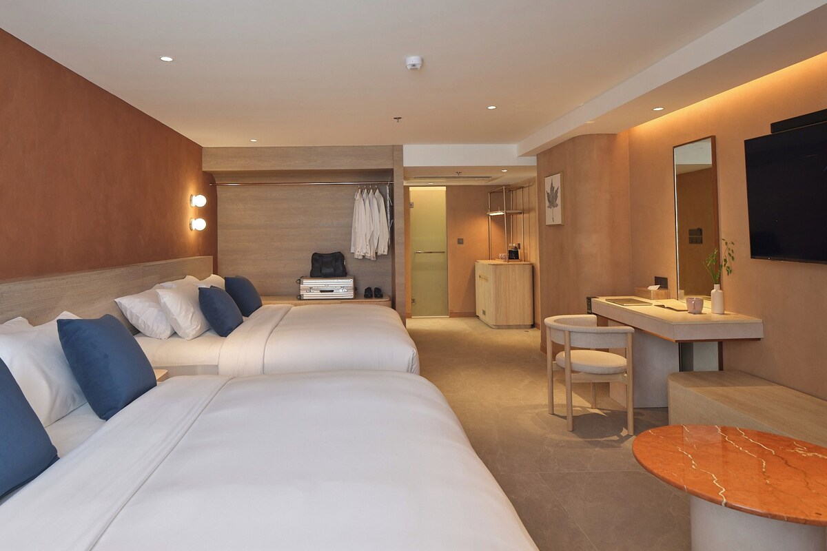 Quarto de hotel com duas camas, área de estar com uma mesa, toucador com espelho e uma pequena cozinha.